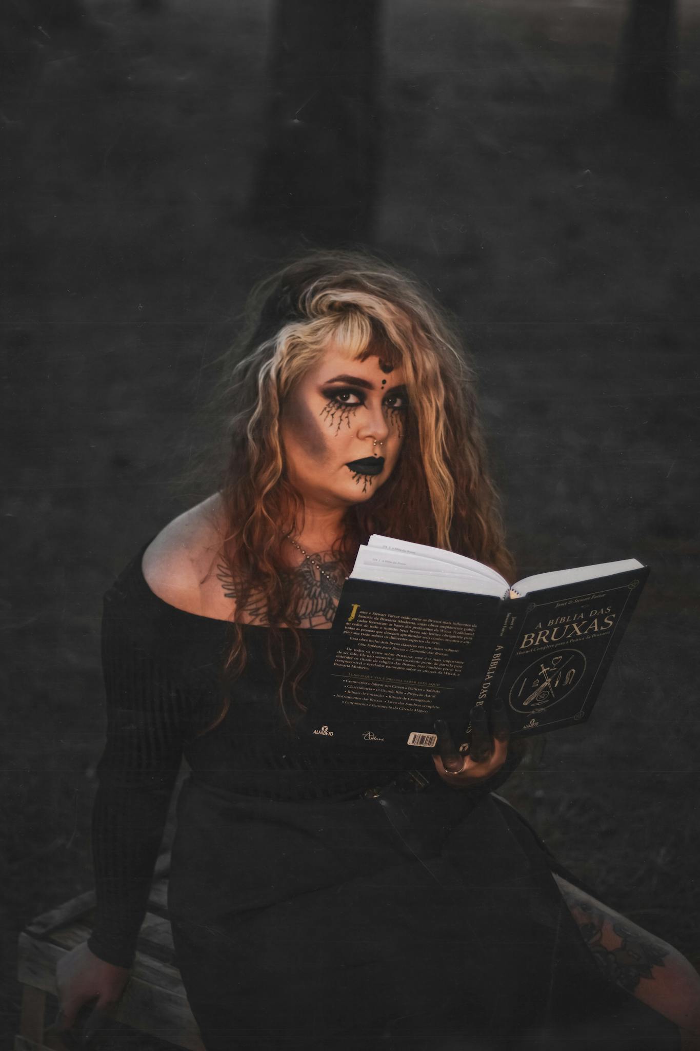A gothic woman dressed as a witch reads a book outdoors in a dark, fantasy forest.