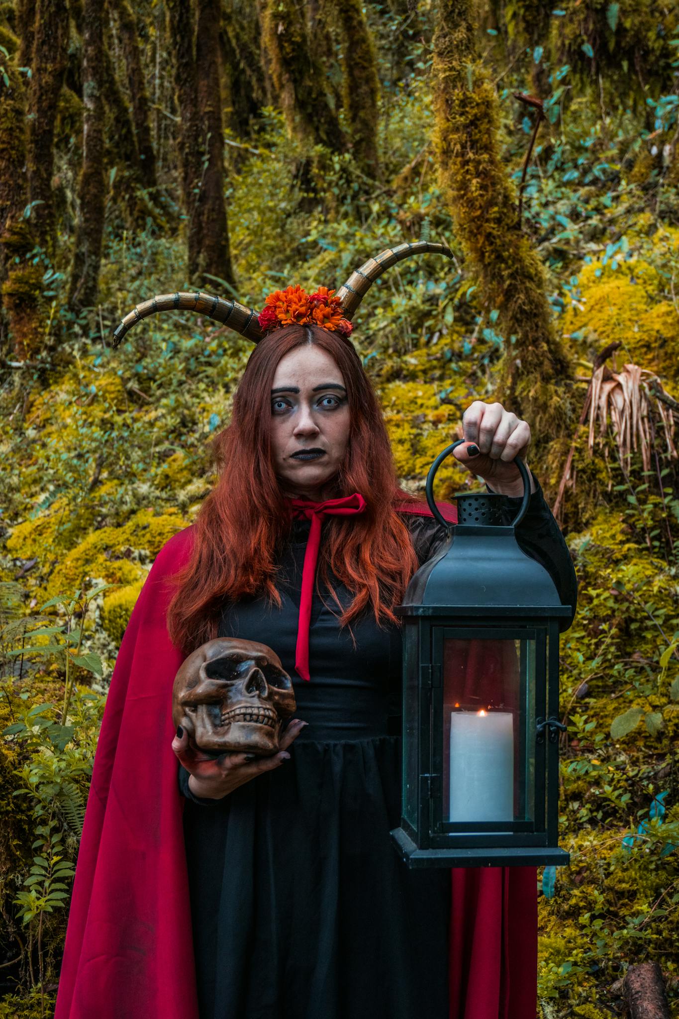 A woman dressed as a witch holding a skull and lantern, standing in a mystical forest.